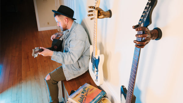 guy in black hat playing guitar while sitting on record player. Next to him are guitars hanging on the wall.
