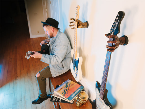 guy in black hat playing guitar while sitting on record player. Next to him are guitars hanging on the wall. 